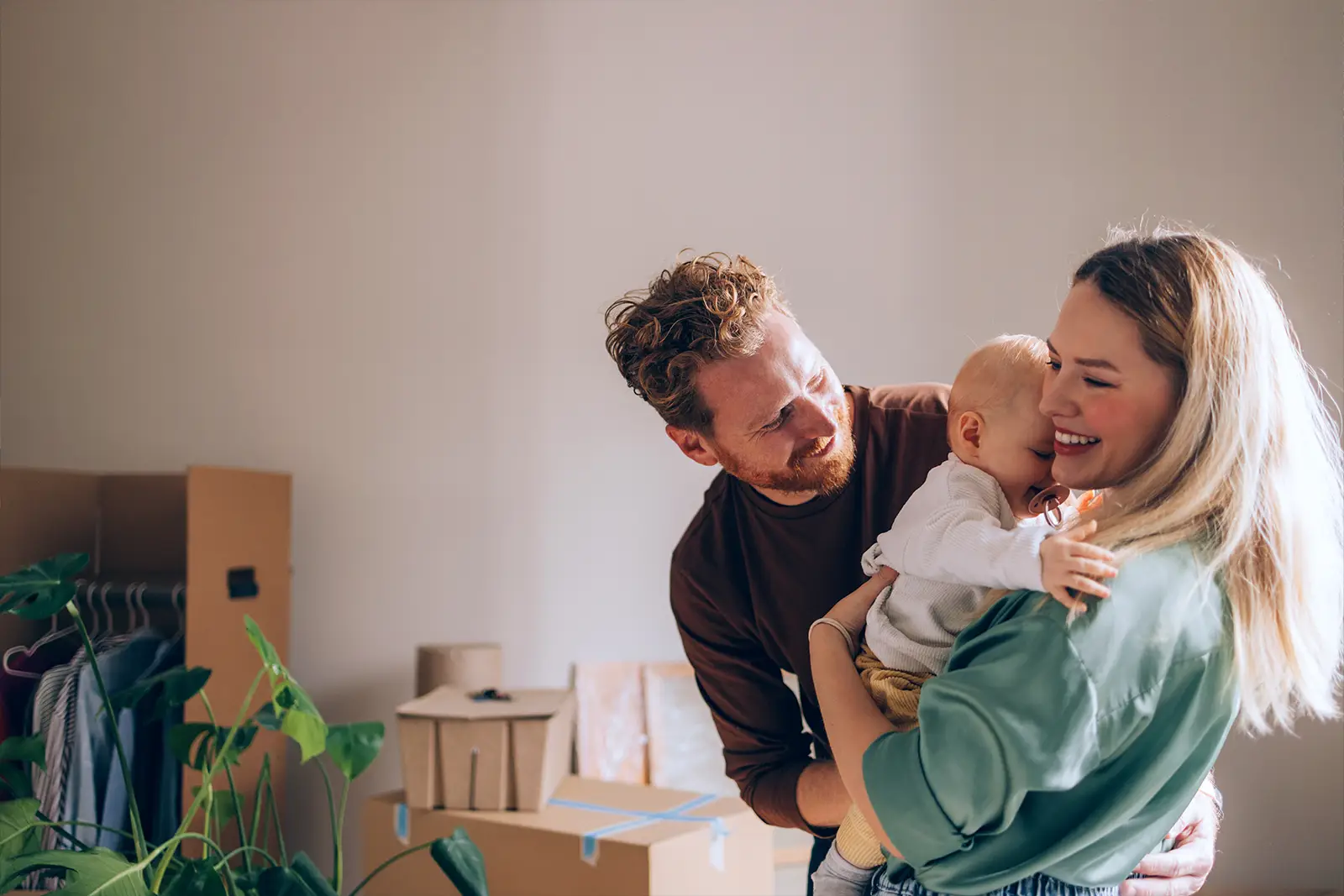Young couple with their baby smiling in their new home with moving boxes in the background.